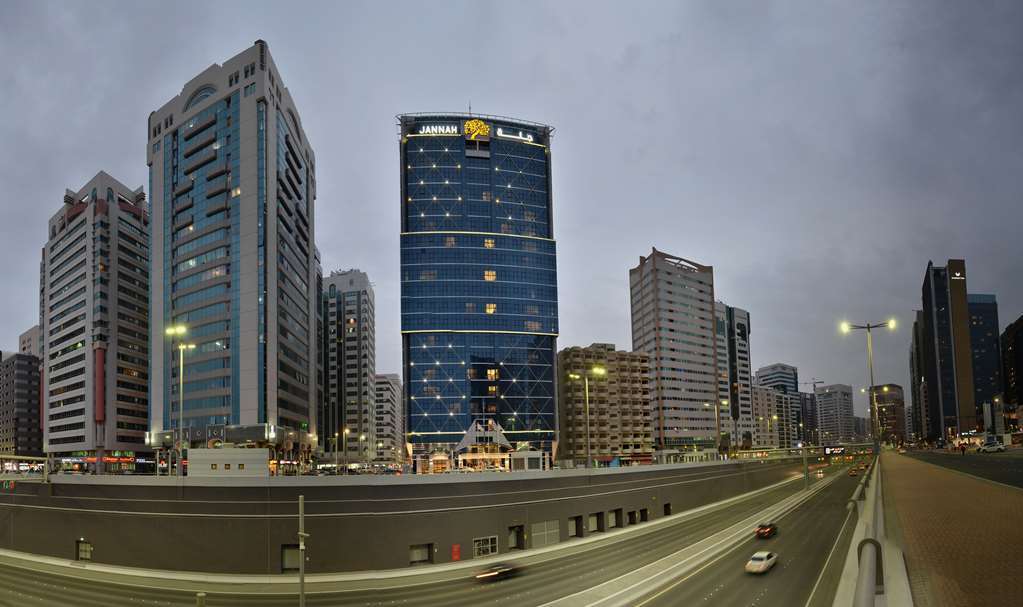 Jannah Burj Al Sarab Hotel Abu Dhabi Exterior foto The photo depicts a modern urban landscape featuring several high-rise buildings. In the foreground, there is a road with vehicles, suggesting a busy city environment. Prominent among the buildings is a tall, blue structure with a distinctive logo at